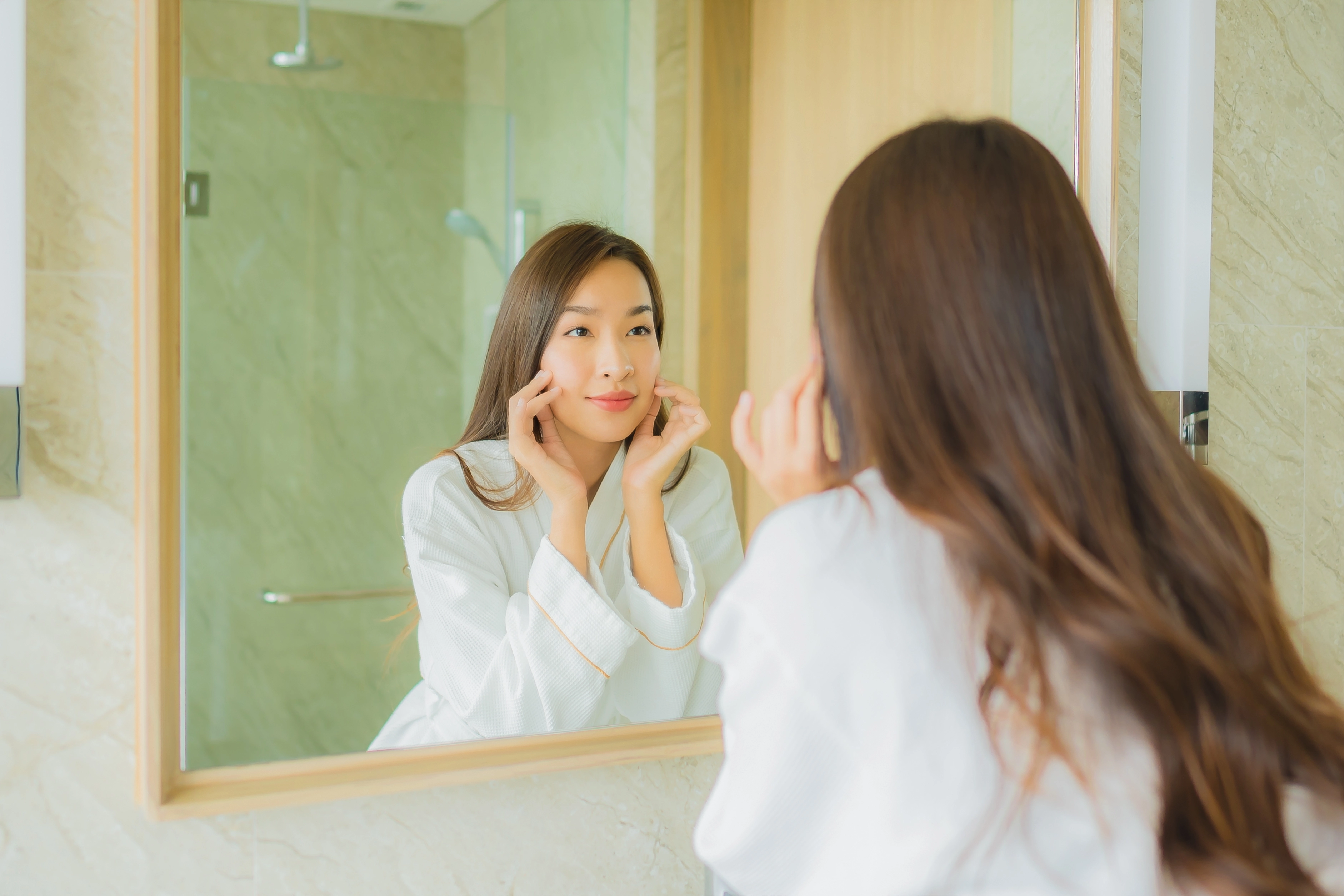 一位年輕美麗的亞洲女性,身穿白色浴袍,在浴室裡對著鏡子檢查自己的臉,看起來很滿意。